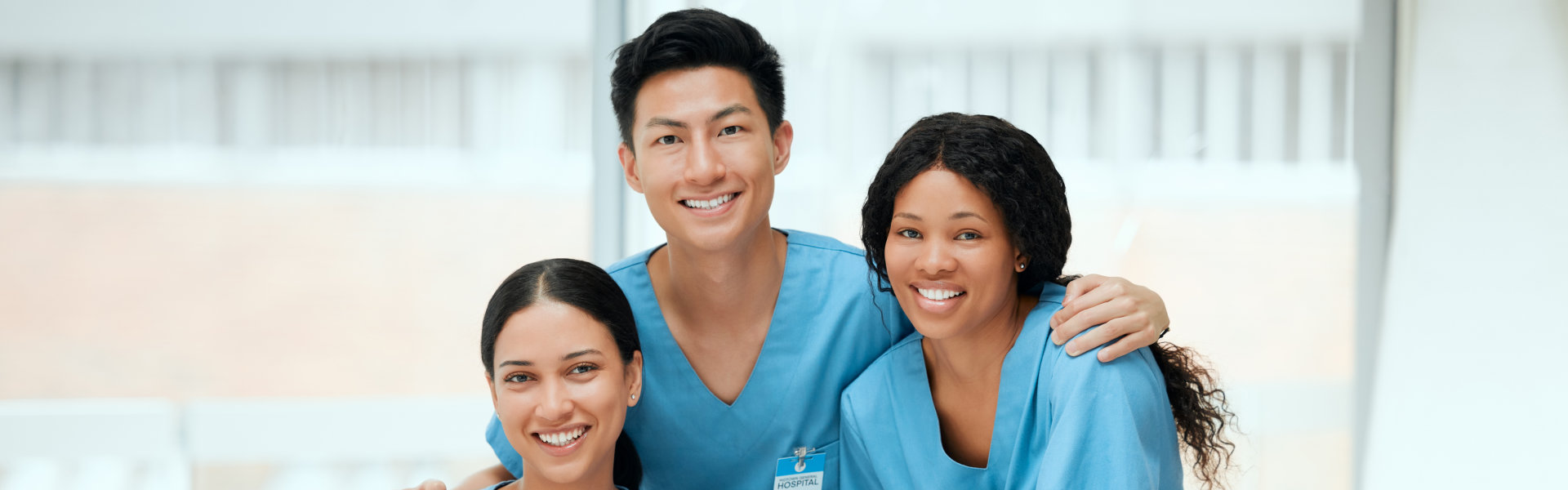 Three individuals wearing blue medical scrubs, standing together with arms around each other, in a bright indoor setting.